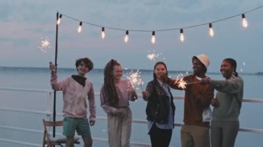 Medium long of multiracial authentic young people smiling, holding sparkling Bengal lights, standing on pier by lake in summer evening