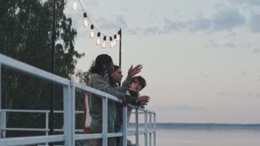 Medium long of young authentic diverse people standing by fencing on lake pier by forest in summer morning, enjoying nature and talking