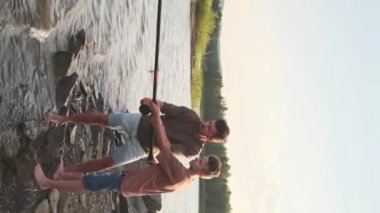 Vertical long of teenage boy and his father wearing shorts and t-shirts, standing on rocky lakeshore on summer day, talking and fishing