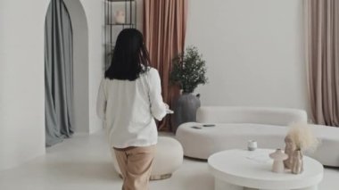 Tracking of Asian businesswoman walking, then sitting on couch in minimalist house, drinking coffee, starting working at daytime