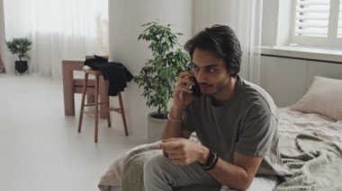 Medium long of young Middle Eastern man in casual clothes sitting on bed in modern apartment, talking on cellphone in morning
