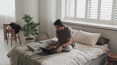 Wide of young Middle Eastern man in loungewear sitting on bed, using laptop computer in minimalist bedroom in morning