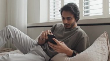 Medium long of young cheerful Middle Eastern man in casual clothes lying on bed, using smartphone in morning, scrolling feed, smiling