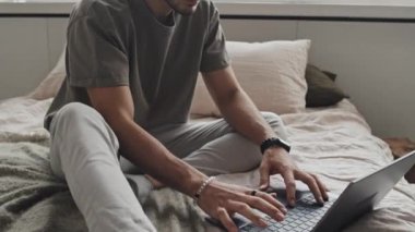 Tilting up of young cheerful Middle Eastern man in loungewear sitting on bed, using portable computer in minimalist bedroom in morning, smiling