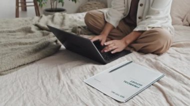 Cropped freelancer in cozy clothing sitting in lotus pose on bed, typing on laptop computer in morning, financial report papers on side