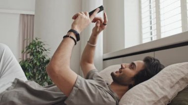 Medium long side-view of young cheerful Middle Eastern man in casual clothes lying on bed, using smartphone in morning, scrolling social media feed, smiling