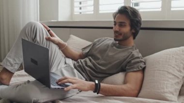 Young cheerful Middle Eastern man in loungewear lying on bed, video calling via portable computer in morning, smiling and talking
