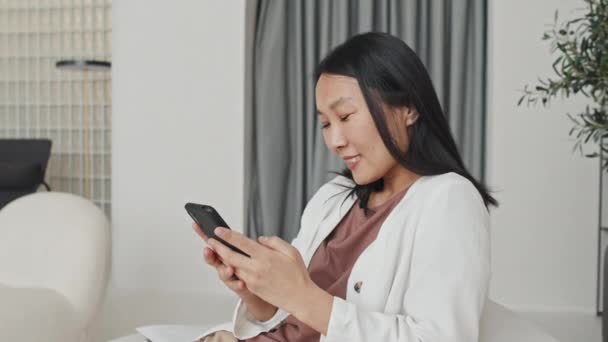 Medium Young Cheerful Asian Woman Typing Smartphone Sitting House Modern — Stockvideo