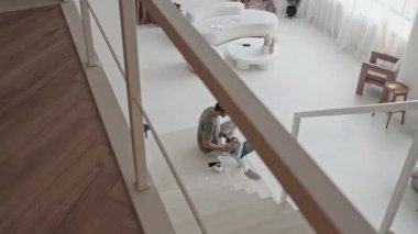 High-angle of Middle Eastern guy sitting on white stairs in house with modern minimalist interior design, using laptop computer and drinking coffee in morning
