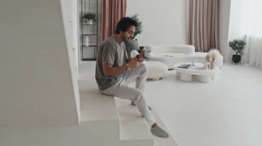 Side view of Middle Eastern guy sitting on white stairs in house with modern minimalist interior design, using smartphone and drinking coffee in morning