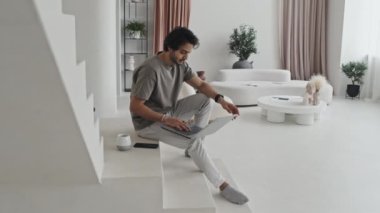 Side-view of Middle Eastern guy sitting on white stairs in house with modern interior design in neutral tones, using portable computer and drinking coffee in morning