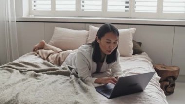 Long shot of young Asian woman in loungewear lying on stomach on bed, typing on portable computer in morning