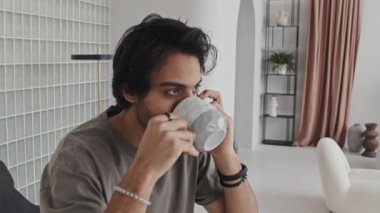 Medium close-up of Middle Eastern young man sitting on in house with modern interior design in neutral tones, talking on cellphone and drinking coffee in morning