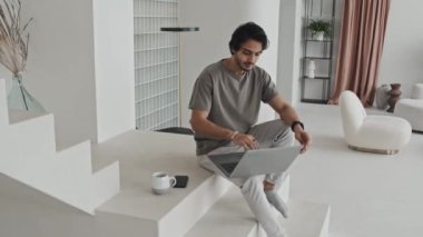 Medium long portrait of Middle Eastern young man sitting on white stairs in house with modern interior design in neutral tones, closing laptop computer, looking and smiling on camera