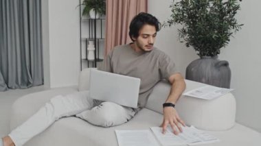 Middle Eastern young man sitting on couch in house with modern interior design in neutral tones, working with documents and portable computer at daytime