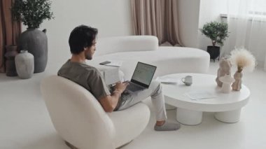 Long over-shoulder of Middle Eastern young male freelancer sitting in living room with modern interior design in muted colors, working with documents and using laptop computer at daytime