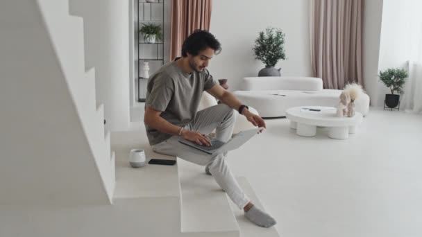 中性調のモダンなインテリアデザインの家の白い階段に座って中東の男のサイドビュー 午前中にポータブルコンピュータと飲料コーヒーを使用してください — ストック動画