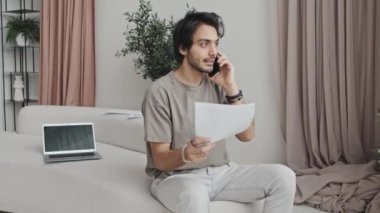 Medium long of Middle Eastern young male entrepreneur sitting on couch in house with modern interior design in neutral tones, holding papers and talking on mobile phone at daytime