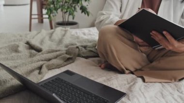 Tilting up of Asian businesswoman in neutral-colored clothes sitting in lotus pose on bed, typing on laptop computer in morning, working with documents