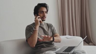Medium long of Middle Eastern young male freelancer sitting in living room with modern interior design in muted colors, using documents and computer, talking on cellphone at daytime