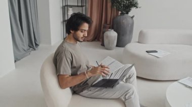 Medium long of Middle Eastern young employee sitting in living room with modern interior design in muted colors, taking notes in notebook and using laptop computer at daytime