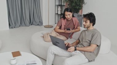 Young Middle Eastern man holding portable computer, asking advice from Asian woman who using smartphone, couple sitting on couch in living room with modern interior design at daytime