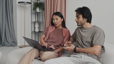 Medium long of young Asian woman using portable computer, Middle Eastern man using smartphone, couple sitting on sofa and talking in living room with minimalist interior design at daytime