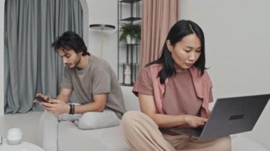 Young Asian woman using portable computer, Middle Eastern man using smartphone, sitting on sofa in living room with modern interior design at daytime