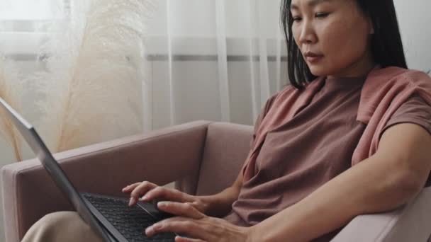 Tilting Young Asian Woman Wearing Muted Colors Sitting Armchair Home — Stok Video