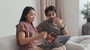Medium long of young Asian woman using portable computer, talking to Middle Eastern man, couple sitting on sofa in living room with minimalist interior design at daytime