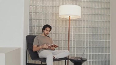 Medium long of Middle Eastern young man sitting in armchair in living room with modern interior design, keeping journal, using laptop in afternoon