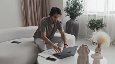 Medium long of Middle Eastern young man sitting on sofa at coffee table, using portable computer, writing in notebook, working with documents in afternoon