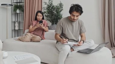 Young Middle Eastern man using portable computer, working with documents, Asian woman using smartphone, sitting on couch in living room with modern interior design at daytime