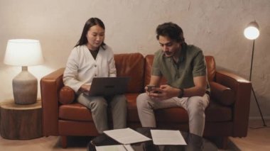 Medium long of two diverse colleagues sitting on brown leather couch in modern coworking space, using portable computer and smartphone in evening