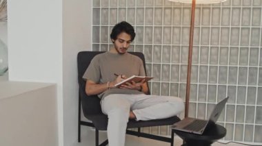 Medium long of Middle Eastern young man sitting in living room with modern interior design, taking notes in notebook and using laptop computer in afternoon