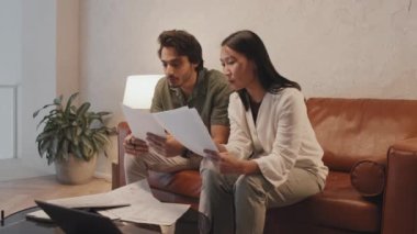 Medium long of two young diverse coworkers sitting on brown leather couch in modern office in evening, having conversation about business papers