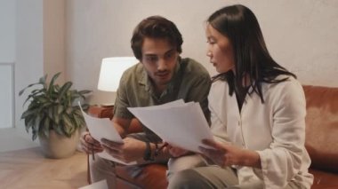 Medium long of two young diverse colleagues sitting on brown couch in lobby of modern office in evening, having conversation about business documents