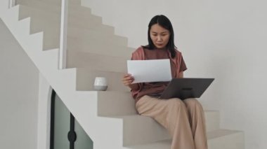 Medium long of young Asian female manager sitting on stairs in modern minimalist office, working with documents, using laptop computer and drinking coffee in afternoon