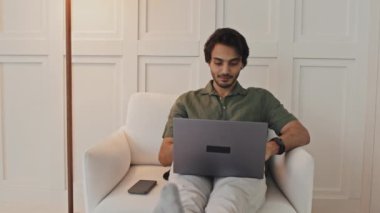 Medium long of young Middle Eastern man sitting in armchair at home, typing on portable computer at daytime