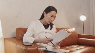 Medium long of successful Asian woman sitting on brown couch at home, doing paperwork in evening