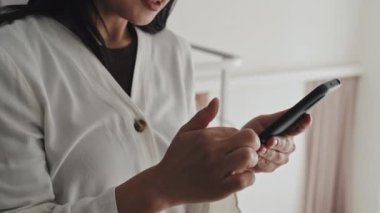 Tilting up of confident young Asian woman standing in house with interior design in light colors, scrolling social media feed on smartphone and smiling at daytime