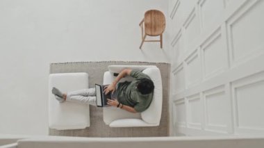 Top view of dark-haired young man sitting in white armchair with legs up on footstool in house with minimalist Scandinavian interior at daytime, typing on laptop computer