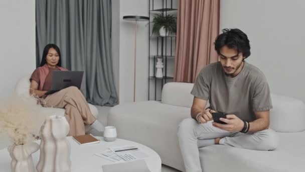 中長い黒髪中東の男性白いソファに座って スマートフォンを使用して アームチェアで若いアジアの女性と話をし 上品なアパートで働いているカップル — ストック動画