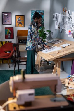 Young creative man standing by wooden table with his artworks and looking for the best drawings for exhibition of modern arts