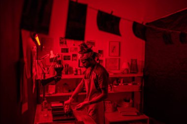 Young modern self-employed photographer pouring photo developer in container while working in dark red lit laboratory