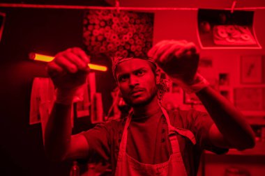 Young serious photographer hanging wet developed black and white photos on thread while working in red lit laboratory