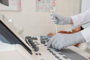 Gloved hands of clinician pushing button of modern ultrasound equipment before medical treatment or examination of patient