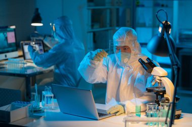 Young contemporary chemist in biohazard suit studying liquid substance while sitting by desk in front of laptop in laboratory