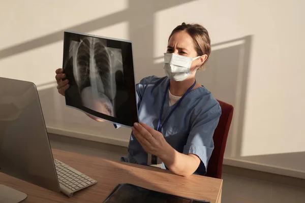 Young female radiologist in protective mask looking at x-ray image of patient while sitting by desk in front of computer