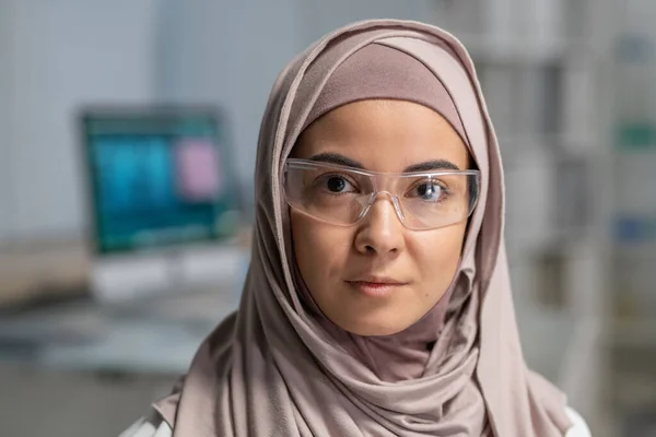 Face of young pretty Muslim female clinician in hijab and protective eyewear looking at camera against her workplace